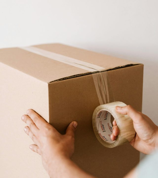 Crop anonymous male using tape to seal packed cardboard box against white wall while moving into new place